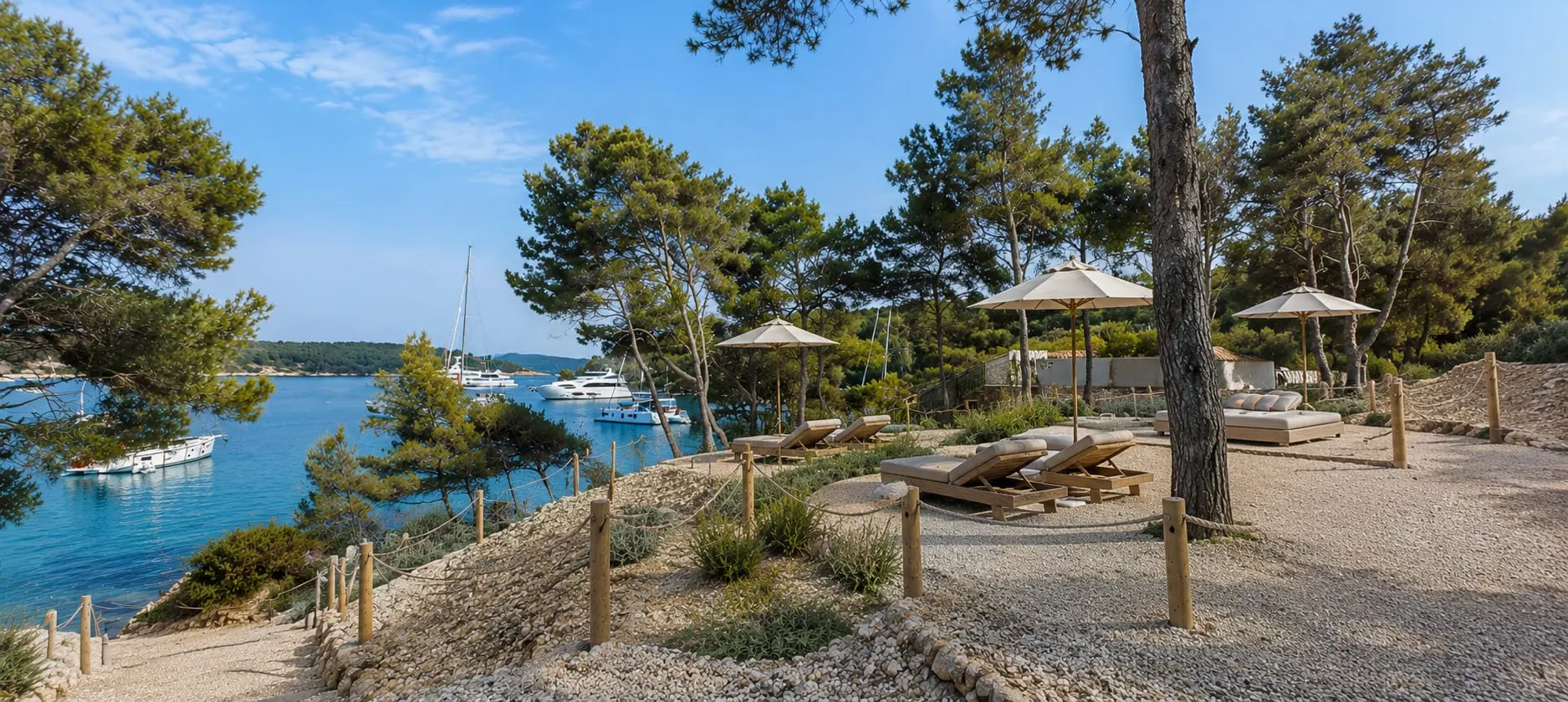 The resort's Beach Park under the pines: loungers and parasols on a gravel terrace above a bay full of anchored boats.
